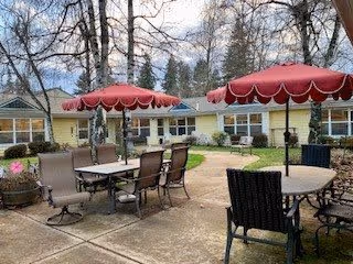 Outdoor patio area with several tables and chairs under red scalloped umbrellas, surrounded by a single-story building with large windows and trees in the background.