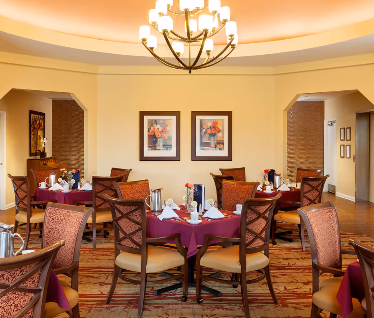 Dining room with round tables covered in burgundy tablecloths, set with white napkins, cups, and silverware. Wooden chairs with patterned upholstery surround the tables. Two framed floral paintings hang on the beige wall, and a large chandelier with multiple lights hangs from the ceiling. The room has a warm and inviting atmosphere.