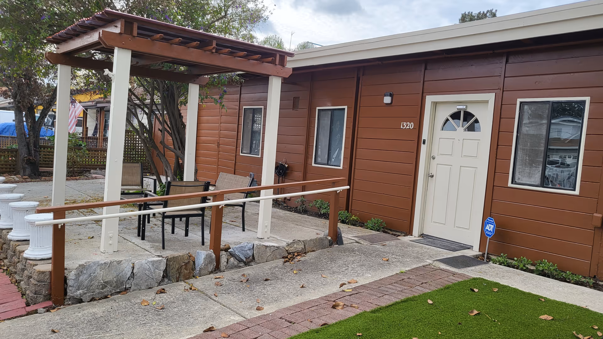 Outdoor patio area with a wooden pergola, several chairs, and a ramp leading to a brown building with a white door and windows. The building has the number 1320 on it and an ADT security sign near the entrance. There are trees and a small lawn area nearby.