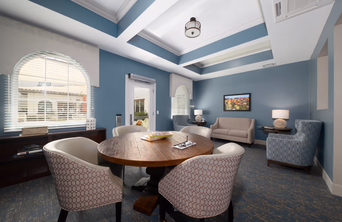 A cozy senior living common area with a round wooden table surrounded by four upholstered chairs. The room has blue walls, two arched windows with white blinds, a beige sofa, two patterned armchairs, two side tables with lamps, and a colorful framed picture on the wall. The ceiling features a tray design with a central light fixture.