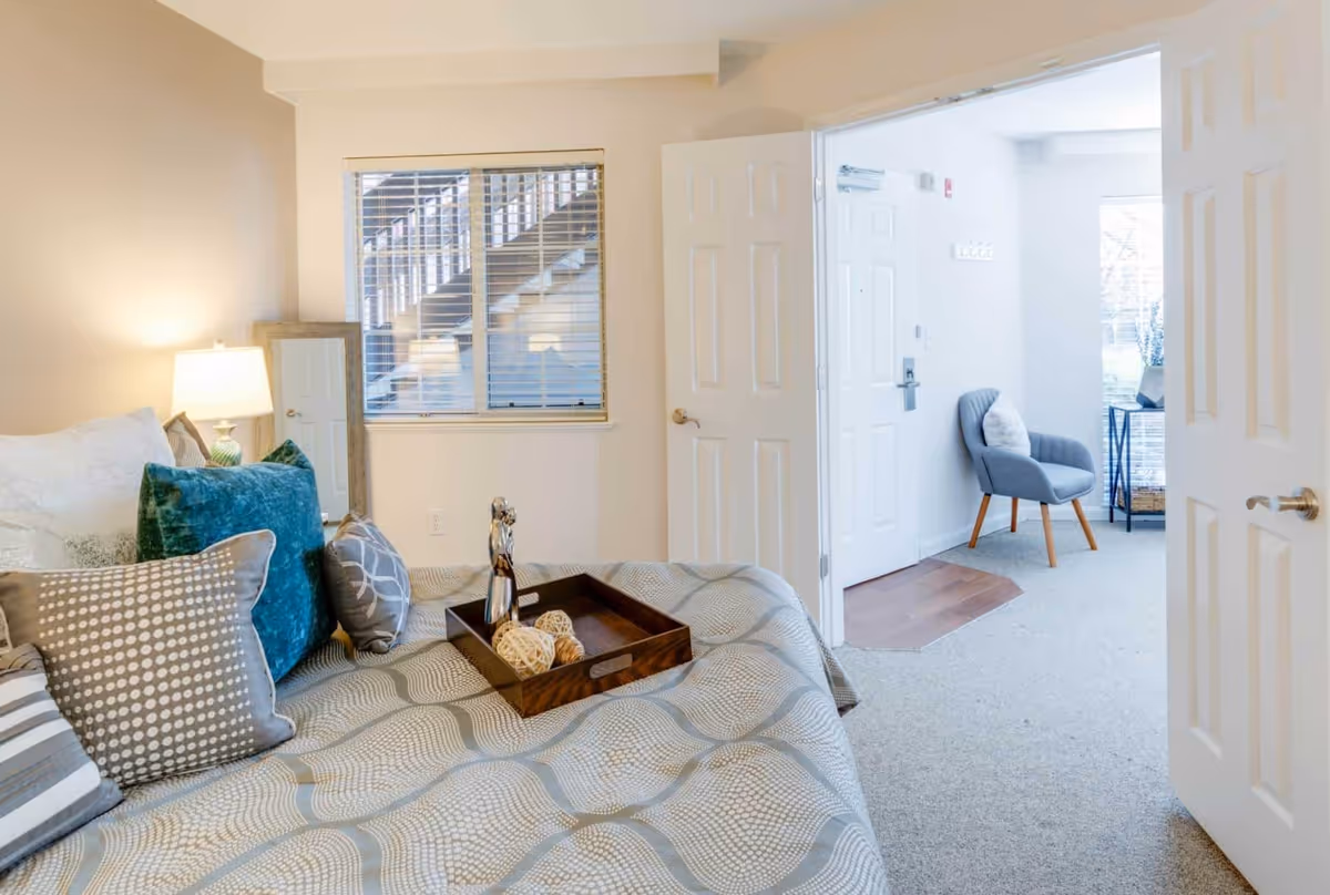 A bright bedroom in a senior living facility with a neatly made bed adorned with multiple decorative pillows and a wooden tray holding decorative items. A bedside lamp is on, and a full-length mirror leans against the wall near a window with blinds. The room opens into a small sitting area with a gray armchair and a side table near a window.