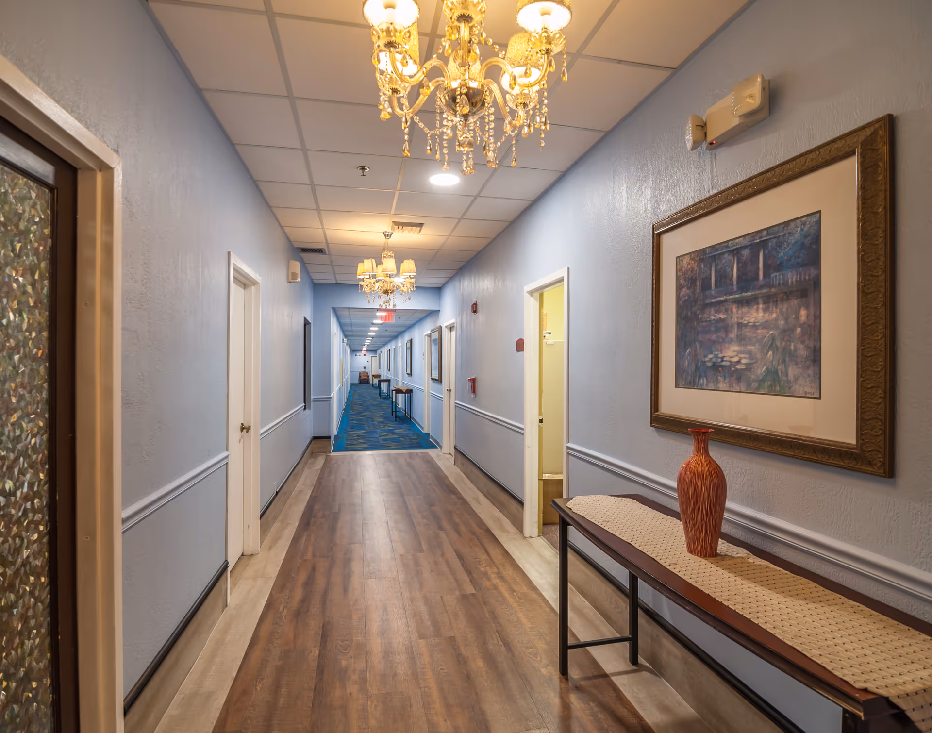 A long, well-lit hallway in a senior living facility with light blue walls, wood flooring, and blue carpet further down. The hallway features multiple doors on both sides, chandeliers hanging from the ceiling, a framed painting on the right wall, and a narrow table with a decorative vase underneath the painting.
