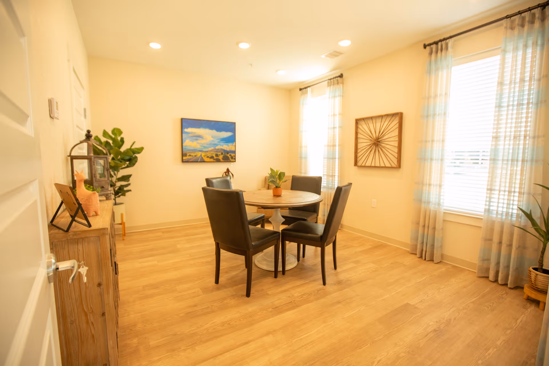Bright dining room with a round wooden table surrounded by four dark chairs, light wood floors, wall art, and two large windows with sheer curtains.