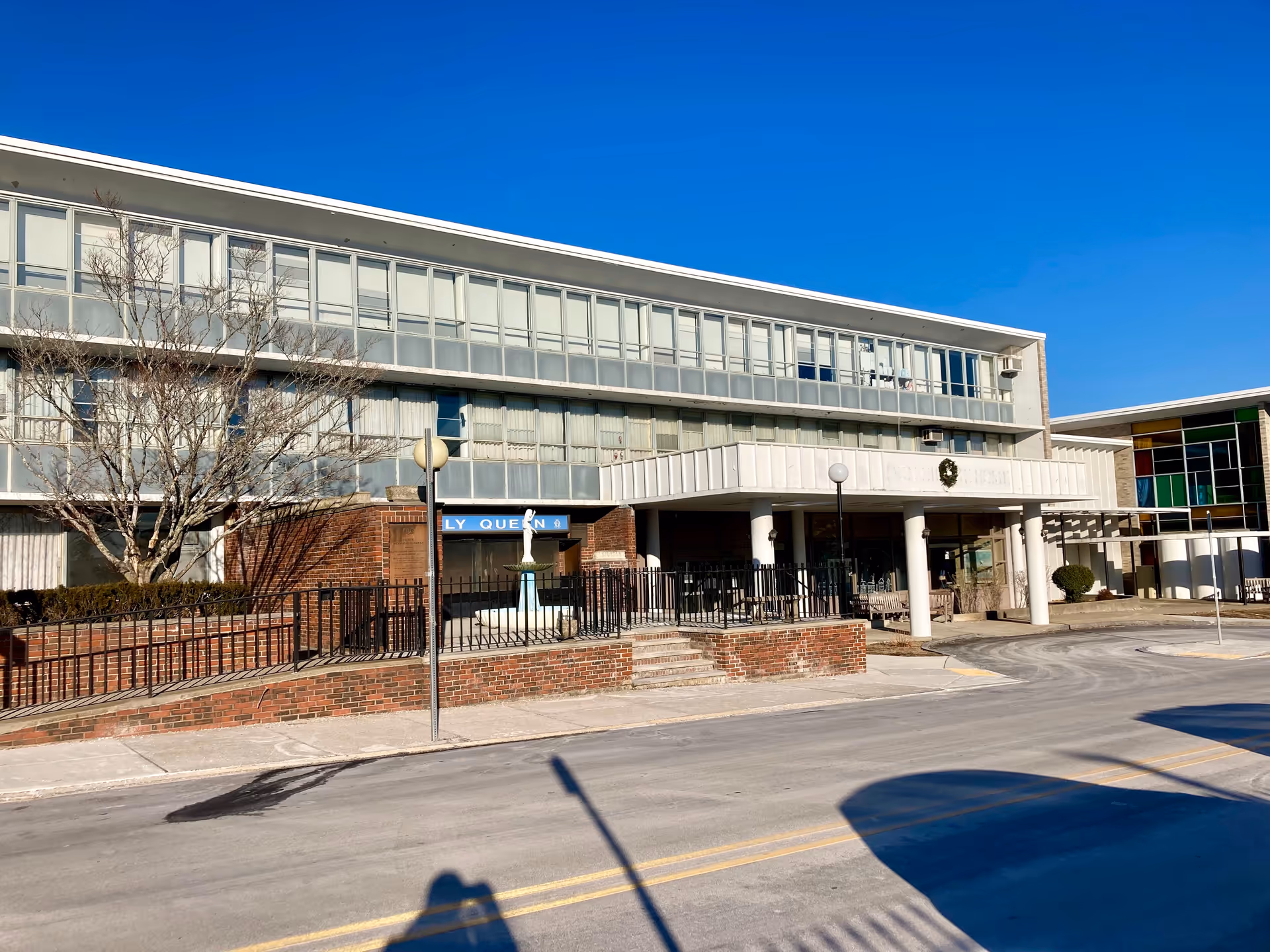 Front exterior of a multi-story senior living building with a covered porte-cochere, brick planters, and a clear blue sky.