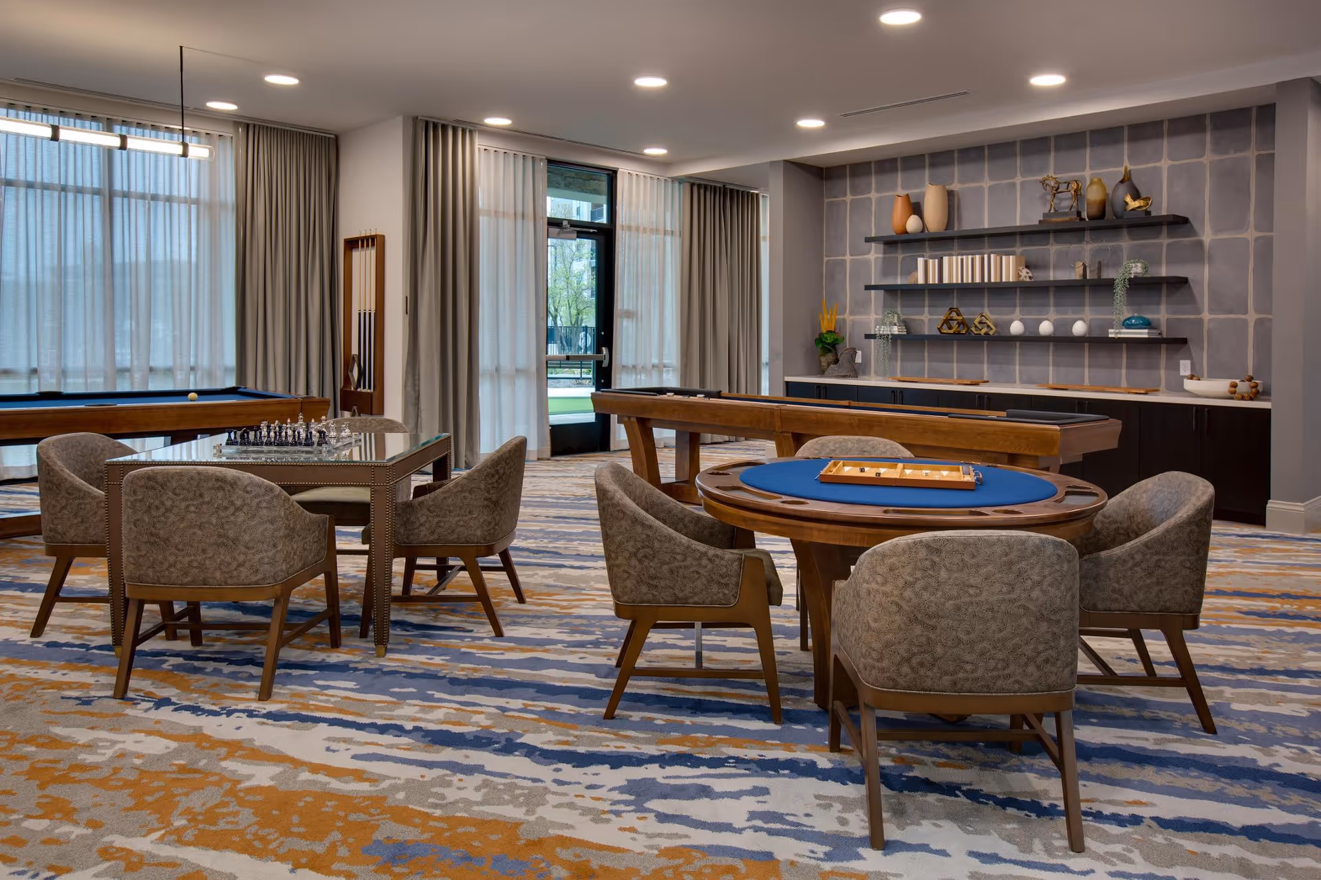 Well-appointed interior game room with a round poker table, a chess table, shuffleboard and seating in front of large windows.