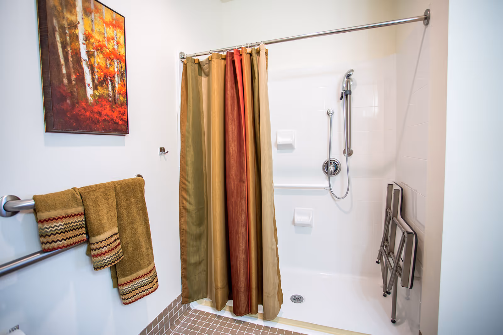 A bathroom shower area with a multicolored shower curtain in shades of green, gold, and red. The shower has white tiled walls, a handheld showerhead, and a foldable shower seat mounted on the wall. On the left wall, there are three brown towels with patterned edges hanging on a towel rack, and a colorful painting of autumn trees is hung above them.
