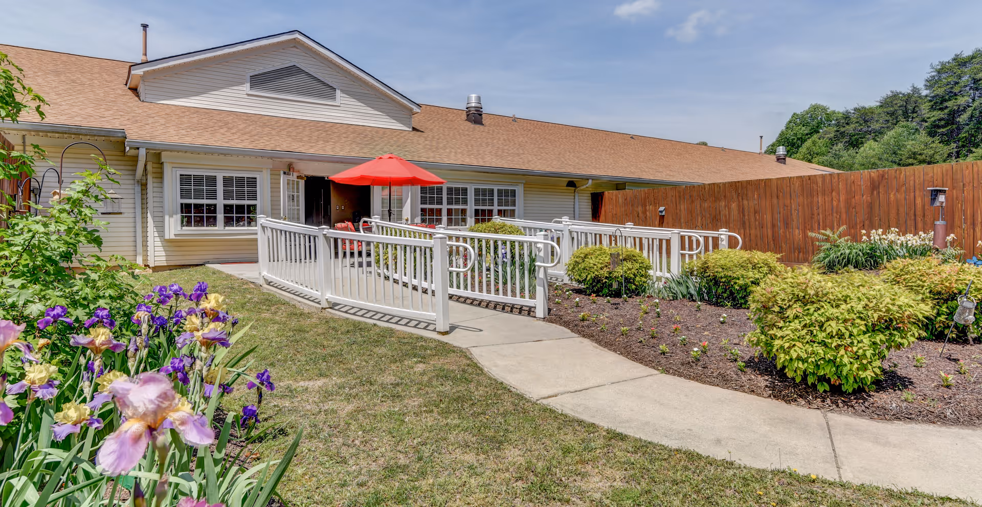 Front exterior of a senior living facility with a wheelchair ramp, patio seating under a red umbrella, and surrounding flower beds.