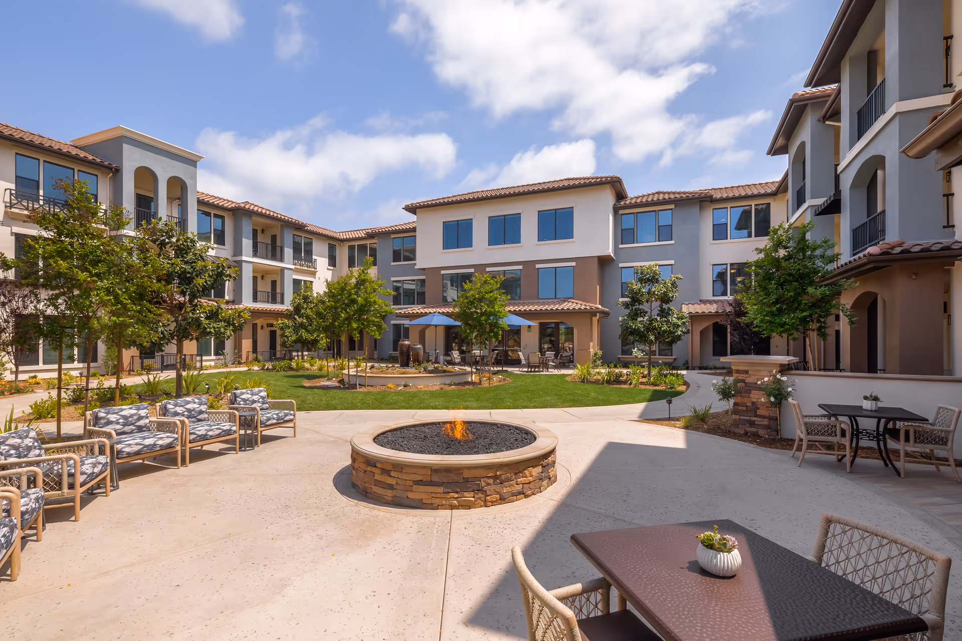 Outdoor courtyard area of a senior living facility with a circular stone fire pit in the center, surrounded by cushioned chairs and tables. The courtyard is landscaped with green grass, small trees, and shrubs, and is enclosed by a three-story building with balconies and large windows under a partly cloudy sky.