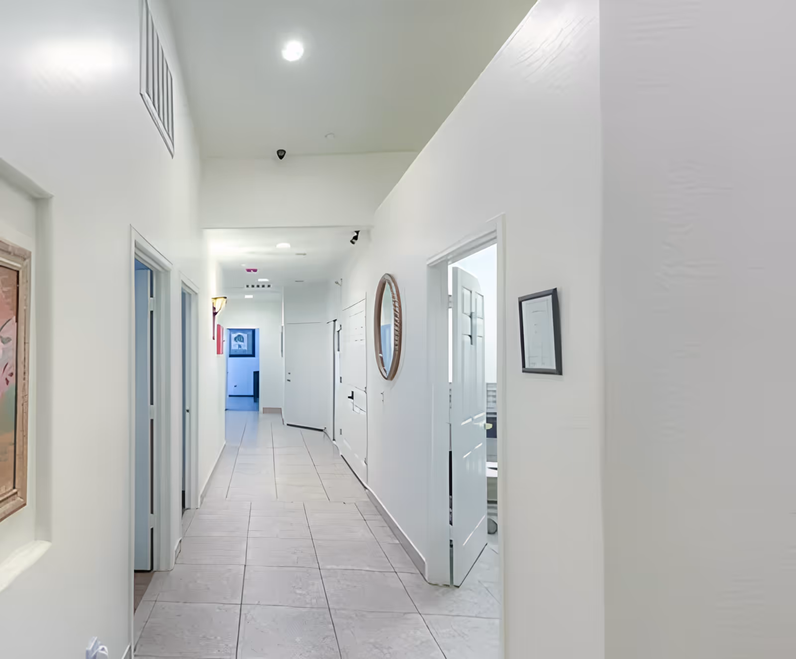 A long, narrow hallway with white walls and tiled floor. Several open doors lead to different rooms, and there are framed pictures and a round mirror hanging on the walls. The hallway is well-lit with ceiling lights.