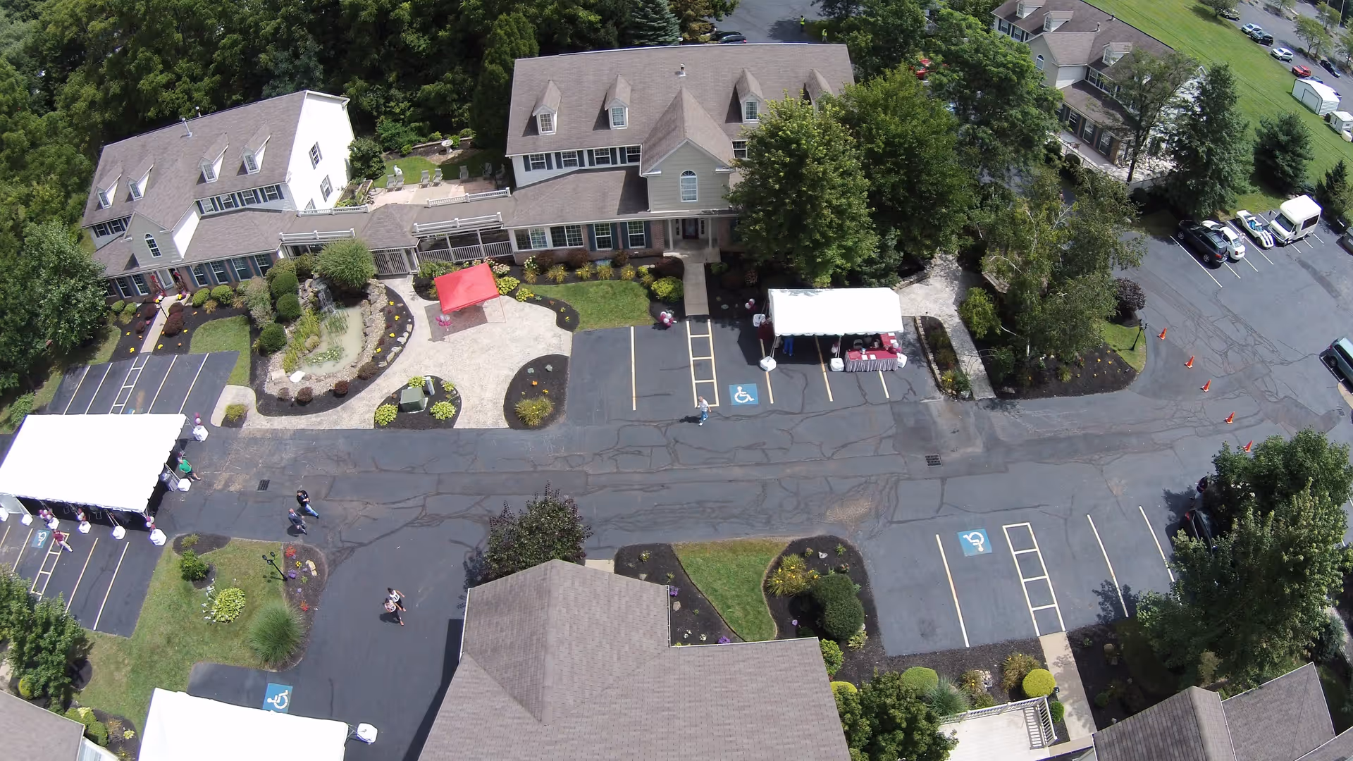 Aerial view of Heritage Villas assisted living facility in Ohio showing multiple connected buildings with pitched roofs, landscaped gardens, paved parking areas with handicap spaces, and several white and red tents set up outside. Trees and greenery surround the property.