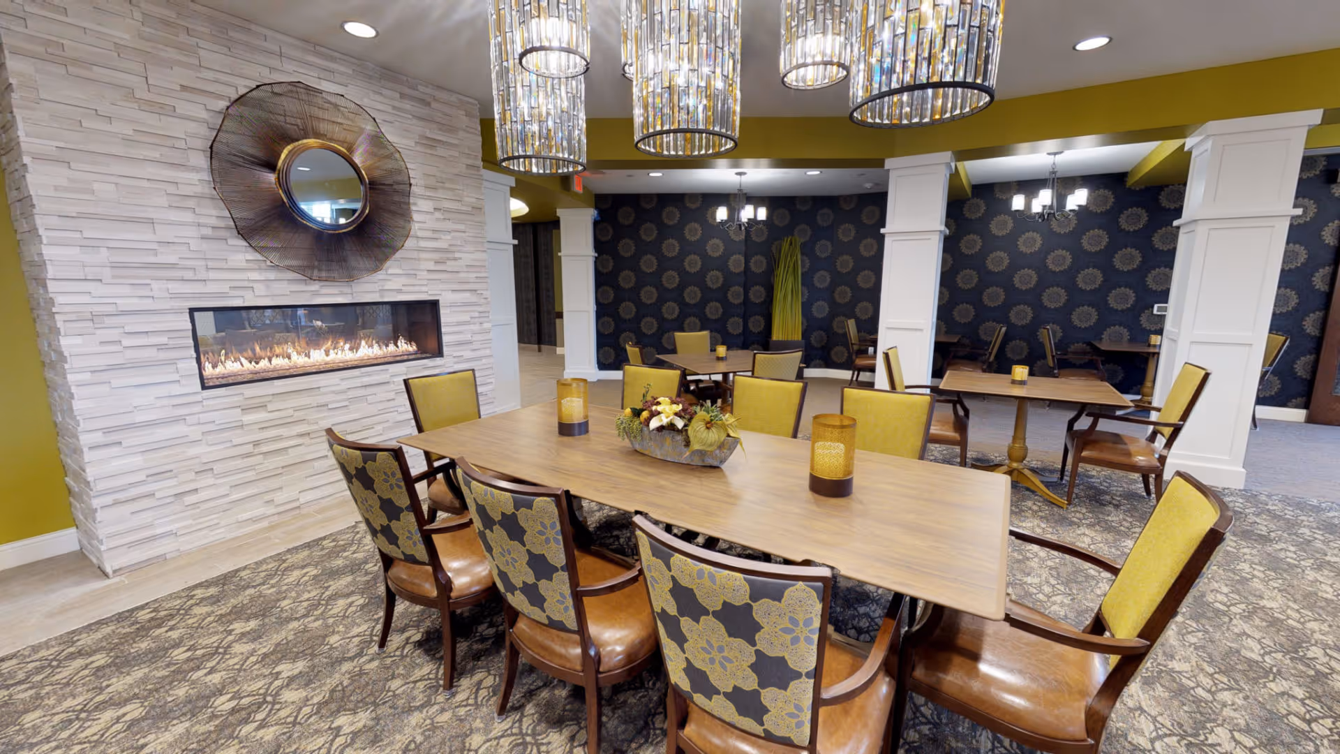 A well-lit dining area in an assisted living facility featuring a long wooden table surrounded by chairs with patterned upholstery and yellow backs. The room has a modern fireplace set in a white textured stone wall with a decorative round mirror above it. Multiple cylindrical pendant lights hang from the ceiling, and additional tables and chairs are visible in the background against a dark wall with a circular pattern.