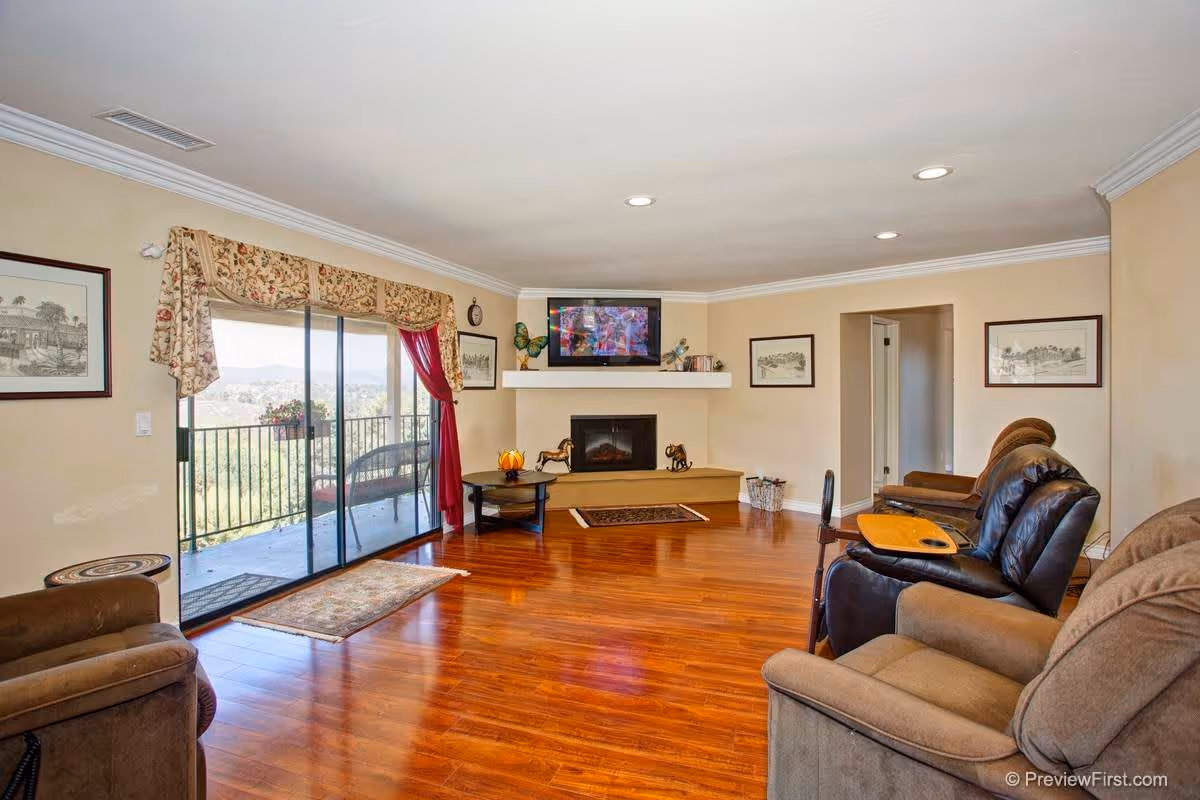 A spacious living room with wooden flooring, beige walls, and recessed ceiling lights. The room features a fireplace with a TV mounted above it, flanked by framed artwork. There are several comfortable recliner chairs arranged around the room, one with a wooden tray attached. A large sliding glass door with floral curtains opens to a balcony with outdoor seating and a scenic view of hills and trees.