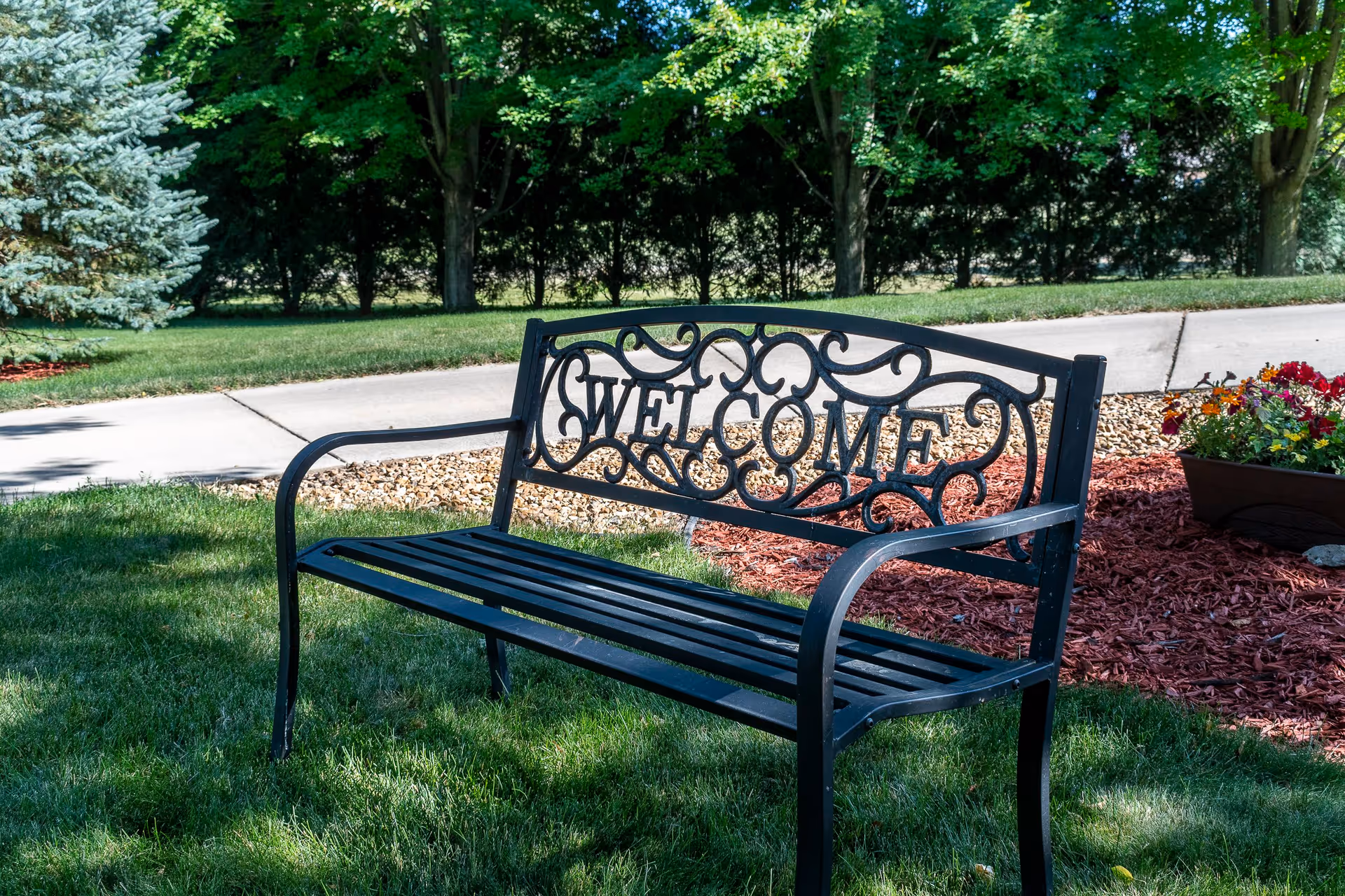 Black metal bench with decorative backrest featuring the word 'WELCOME' in a garden area with green grass, trees, a sidewalk, and a flower planter with colorful flowers.
