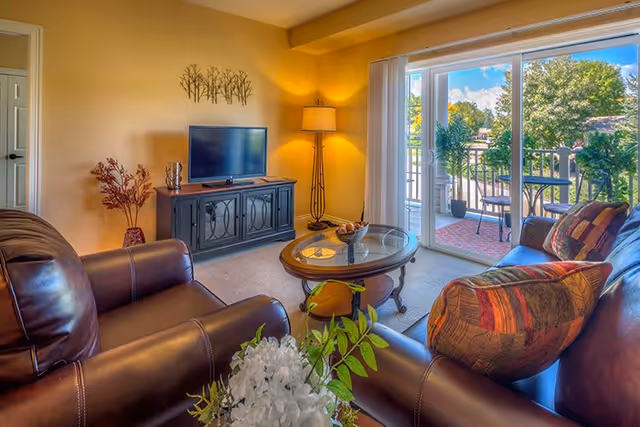 A cozy living room with leather armchairs and a sofa with patterned cushions. A glass coffee table is in the center, and a TV sits on a dark wooden cabinet against a beige wall. A tall floor lamp is lit in the corner, and sliding glass doors open to a balcony with outdoor seating and a view of trees and a clear sky.