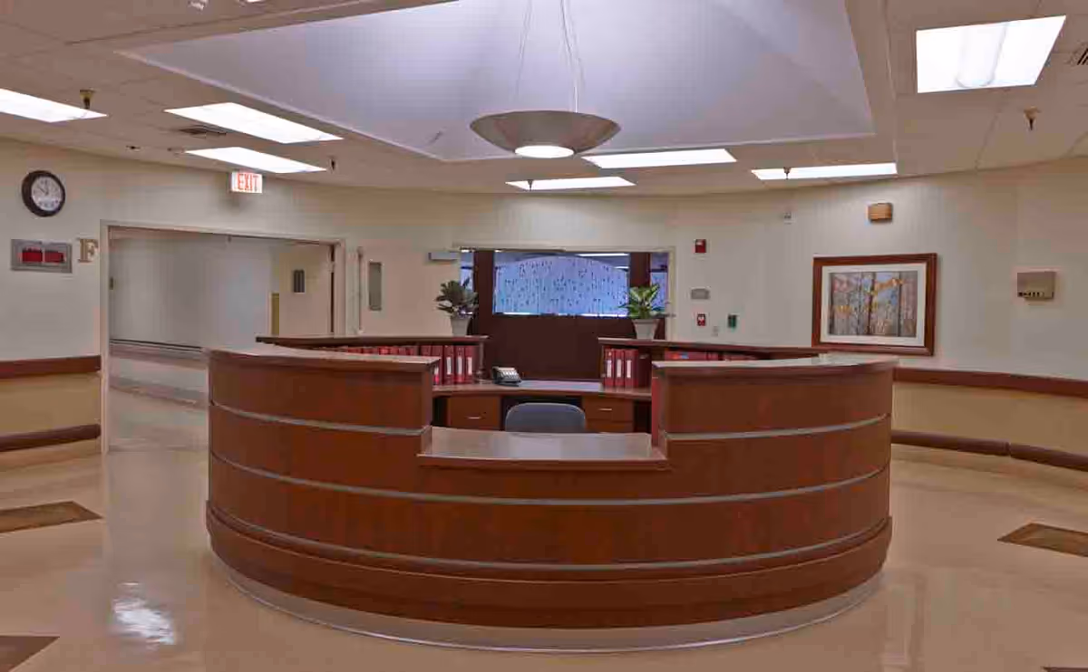 A curved wooden reception desk in a hallway of a senior living facility with beige walls, a clock on the wall, an exit sign, and a framed picture. The desk has office supplies and plants on it, and the area is well-lit with ceiling lights.
