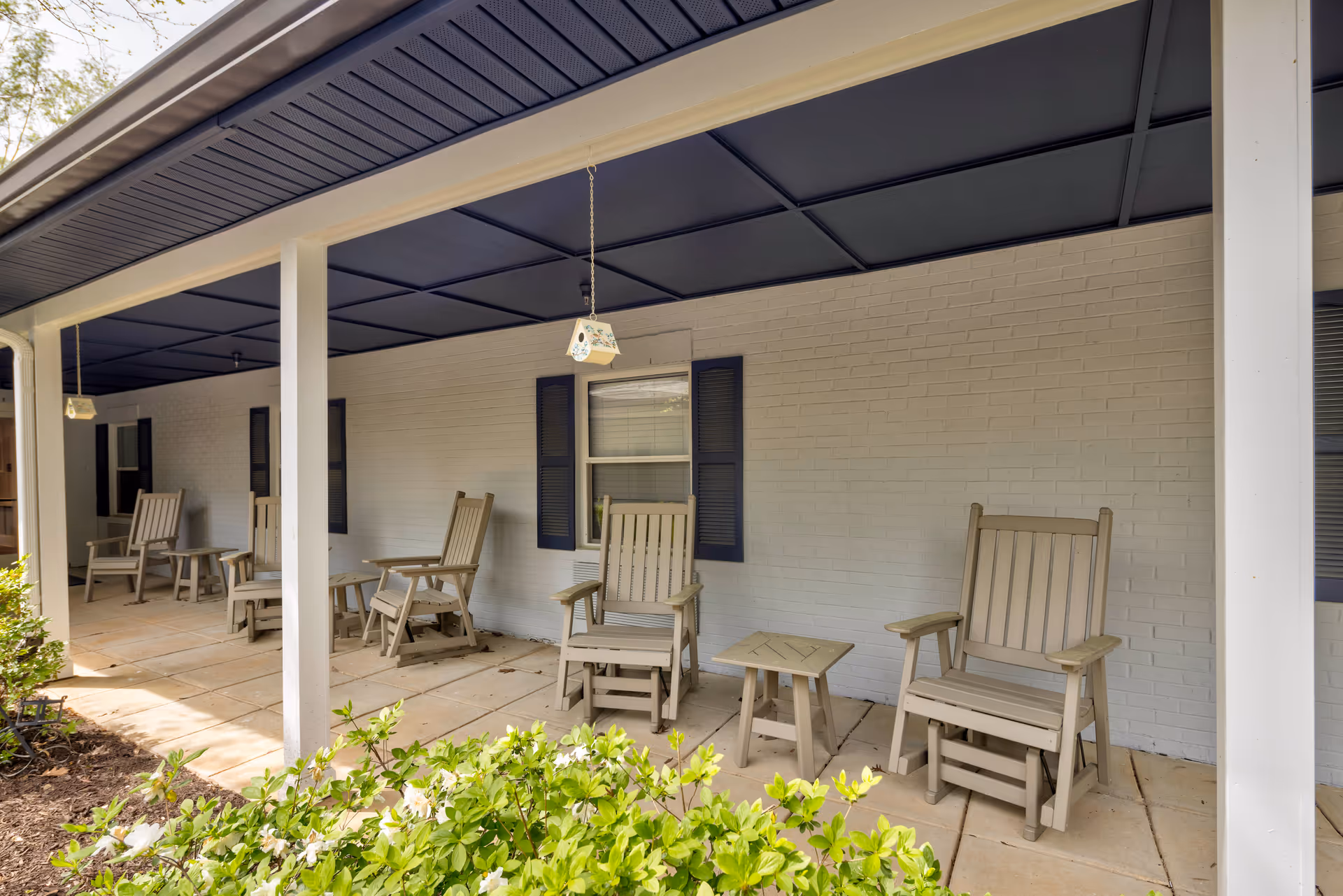 Covered outdoor patio area with several beige wooden rocking chairs and small tables arranged along a white brick wall with windows that have dark blue shutters. There are hanging birdhouses from the ceiling and green bushes in the foreground.