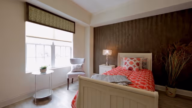 A bright bedroom with a white single bed dressed in red patterned bedding, a nightstand and lamp, a chair by a large window, and a potted plant.