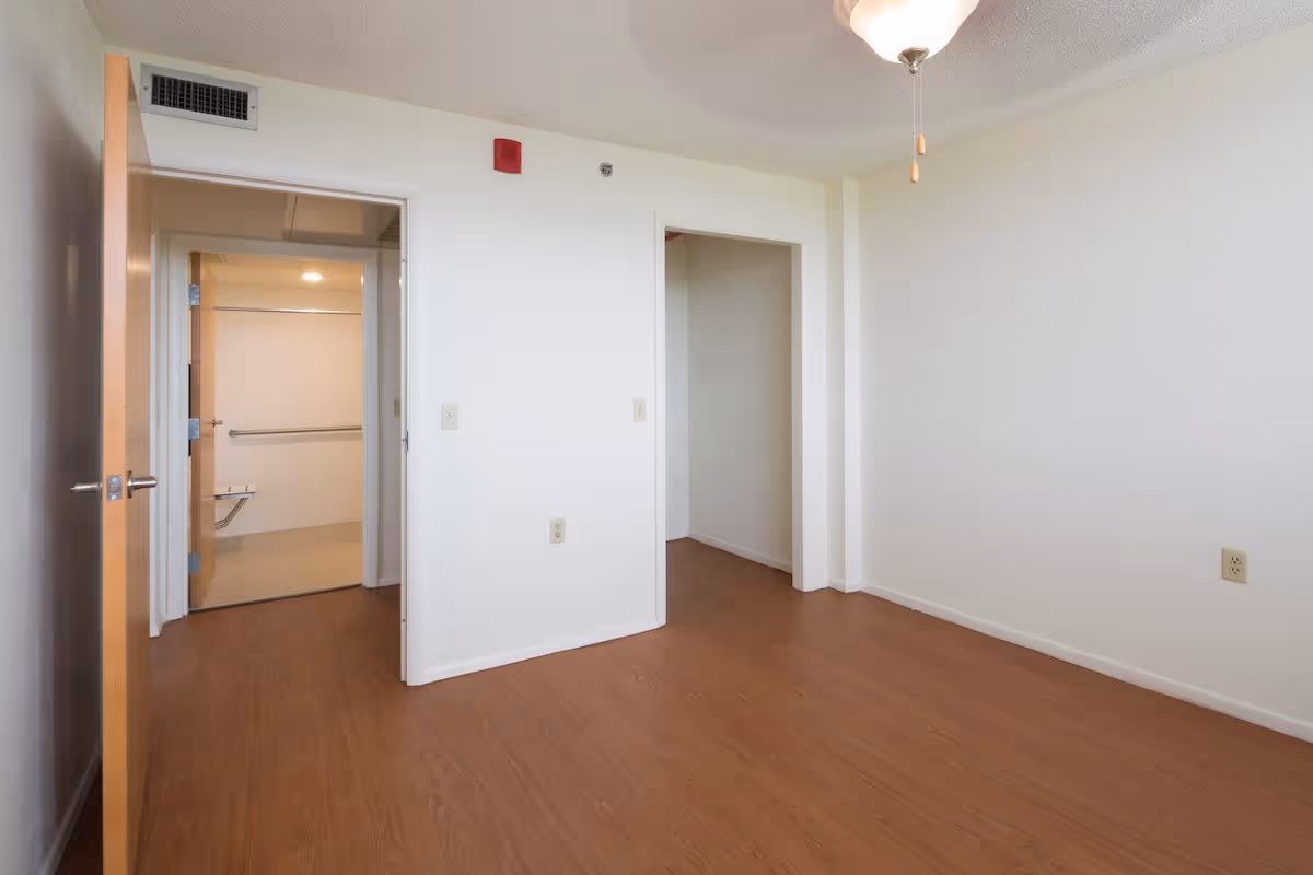 Empty room with wood floors and white walls, a ceiling fan, and open doorways to a bathroom and closet.