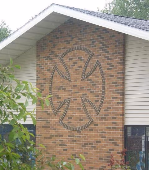Exterior view of a house gable with a tall brick panel featuring a circular decorative brick pattern, vinyl siding, roof eaves, and nearby foliage.
