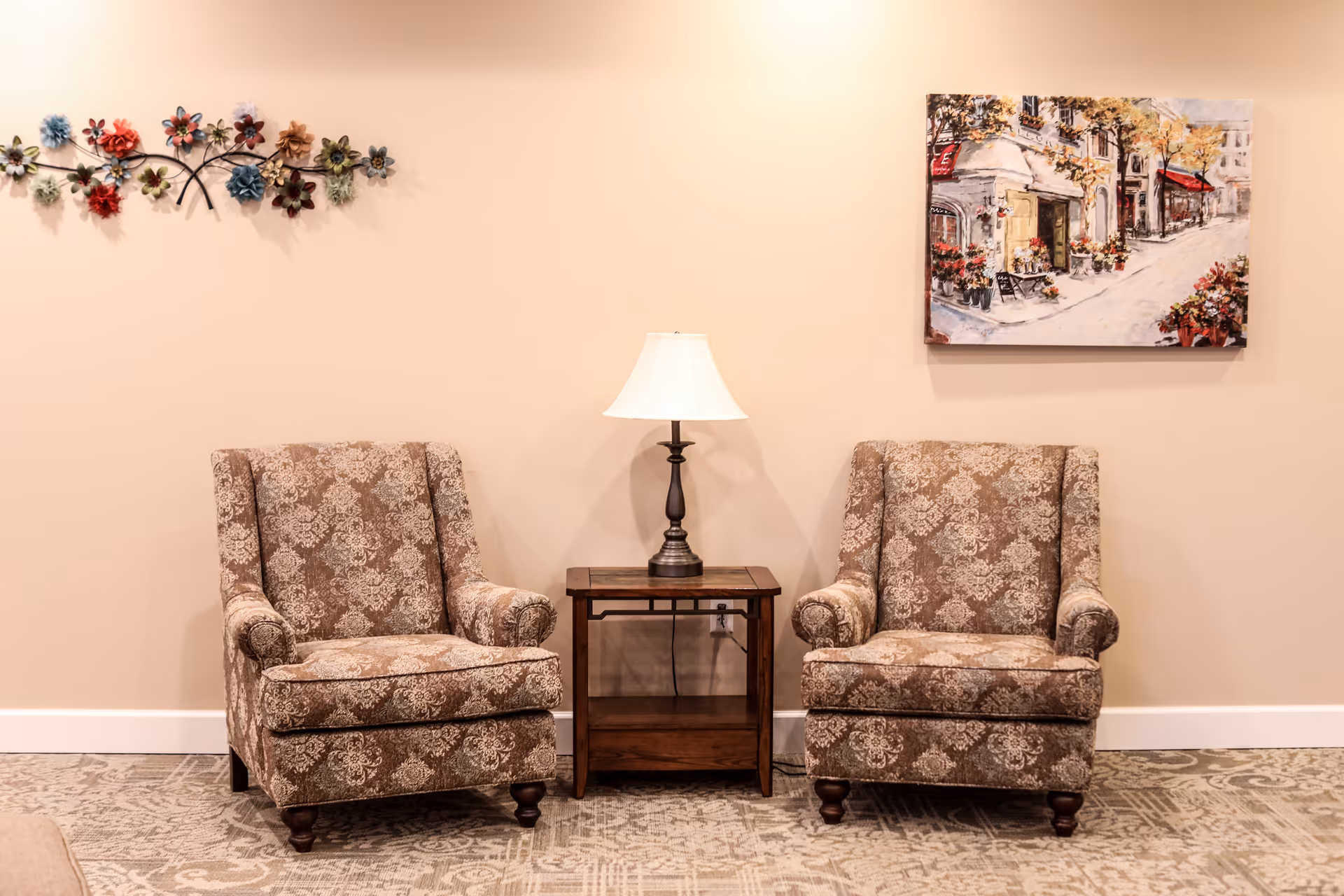 Two patterned armchairs with rolled arms are positioned on either side of a wooden side table with a lamp on it, against a beige wall. Above the left chair is a decorative metal wall art with colorful flowers, and above the right chair is a framed painting of a street scene with buildings and flowers.