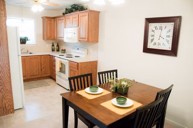 A small kitchen and dining area with wooden cabinets, a white stove, microwave, and refrigerator. The dining table has four black chairs and is set with two place settings including green bowls and plates on yellow placemats. A large wall clock with Roman numerals hangs on the wall above the table.