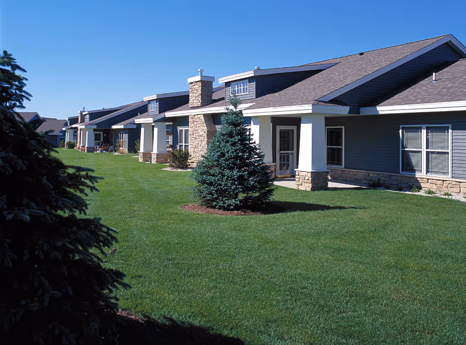 Exterior view of a single-story residential building with stone and siding facade, multiple windows, and a well-maintained green lawn with small evergreen trees under a clear blue sky.