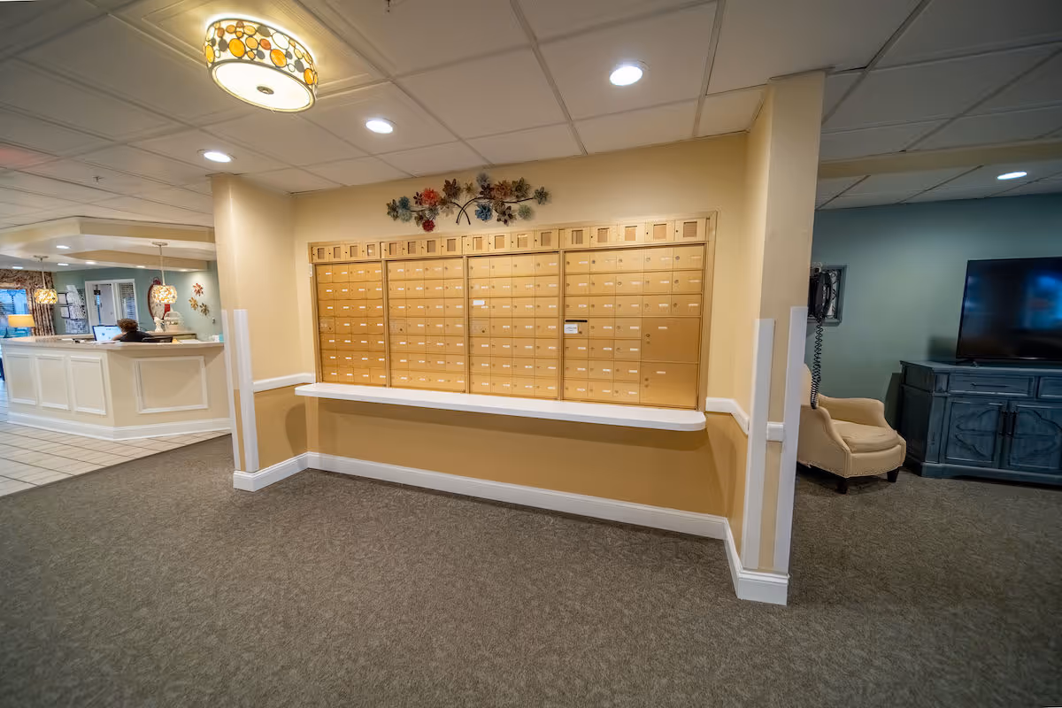 Interior view of a senior living facility showing a wall with multiple mailboxes. To the left, there is a reception desk with a person sitting behind it, and to the right, a seating area with a beige armchair and a television on a blue cabinet. The ceiling has recessed lighting and a decorative ceiling light fixture.