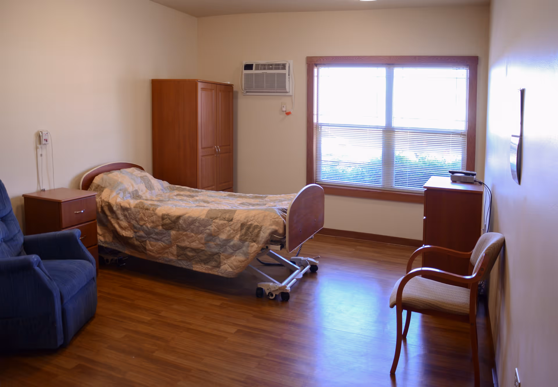 Simple senior living bedroom with an adjustable bed, wooden furniture, a recliner and a window with blinds.