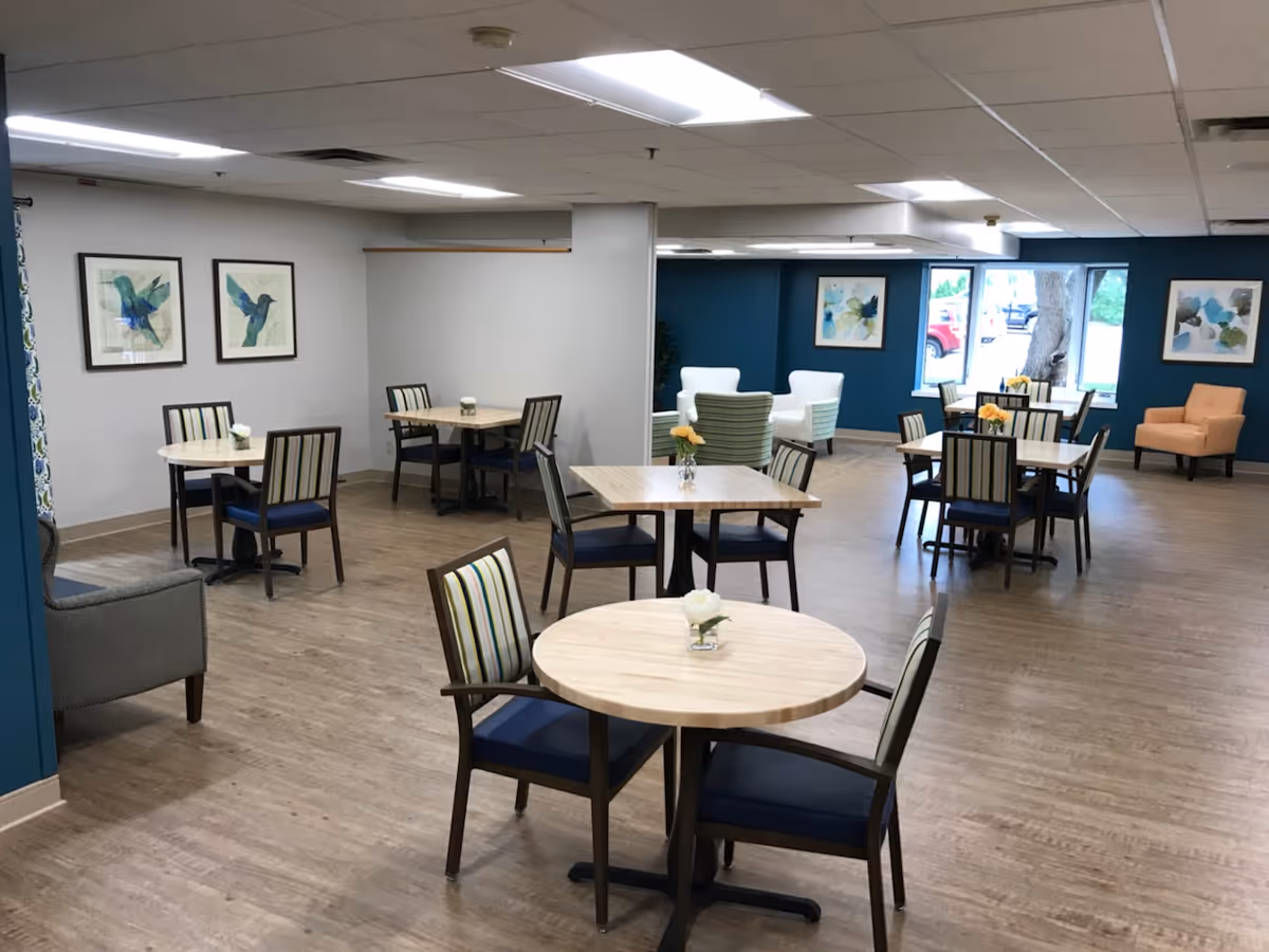 A spacious dining area with multiple round and square wooden tables, each surrounded by chairs with striped upholstery. The room has light wood flooring, white walls with framed bird artwork, and a blue accent wall with additional framed artwork. There are also a few armchairs near the windows, which let in natural light.