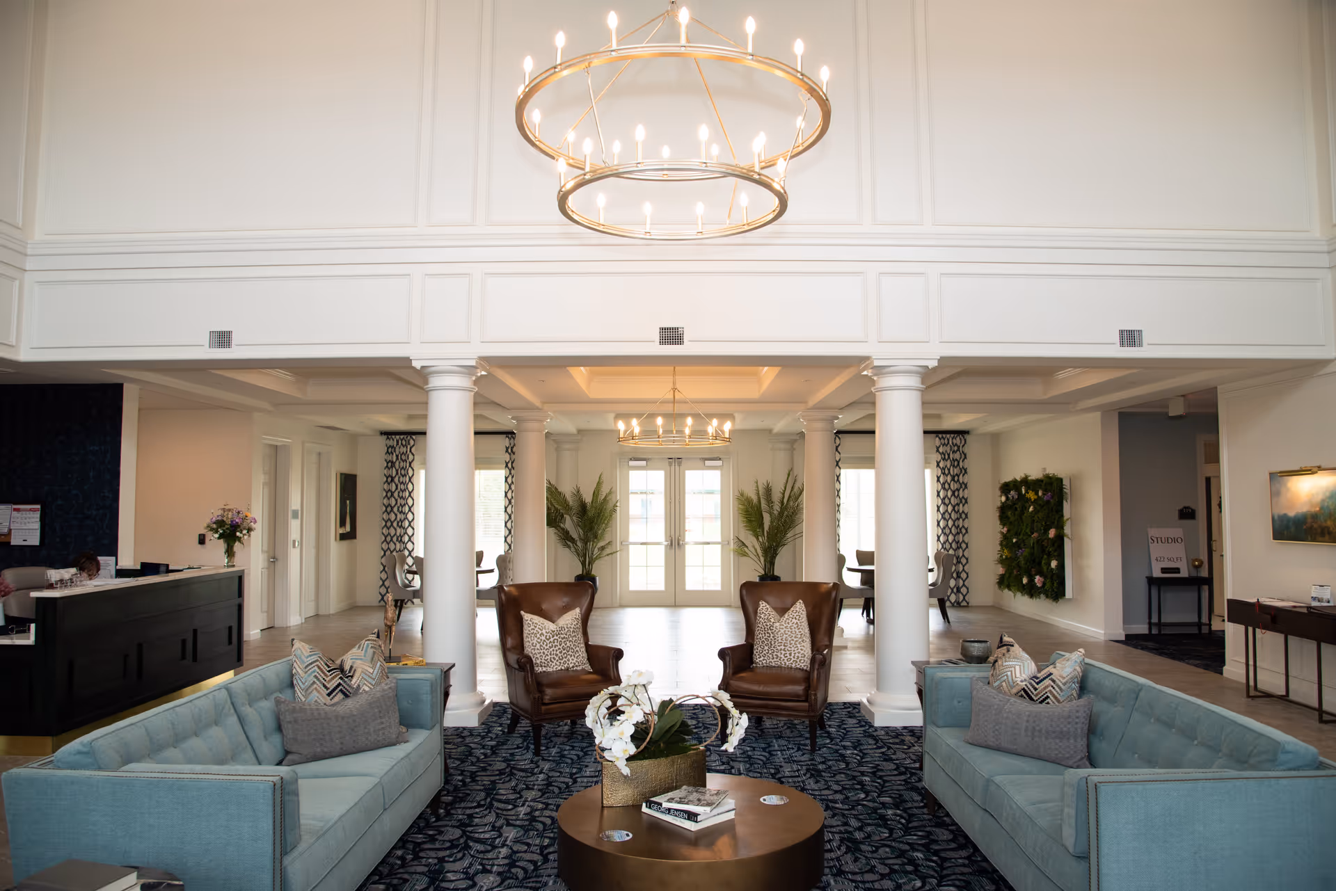A spacious and elegant living room area with two light blue sofas facing each other, two brown leather armchairs with patterned pillows, a round coffee table with a flower arrangement and books, white columns, large windows with patterned curtains, and modern circular chandeliers hanging from a high ceiling.