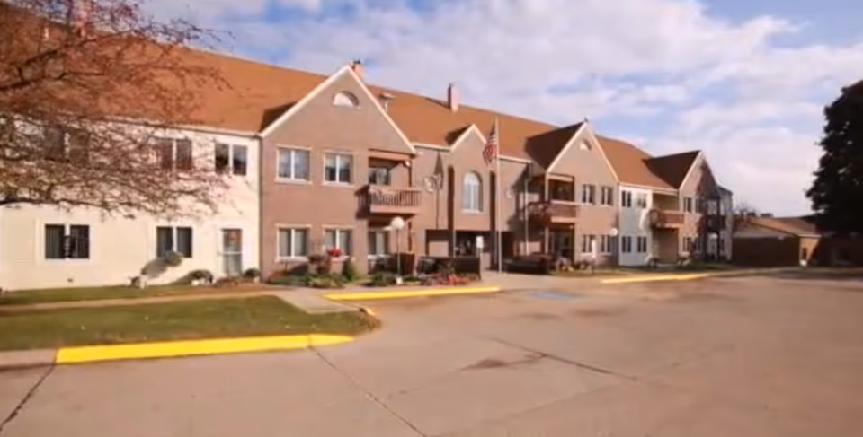 Exterior view of a two-story senior living community building with a mix of brick and siding facade, balconies, and an American flag near the entrance. The building is surrounded by a paved driveway and some landscaping with grass and trees.