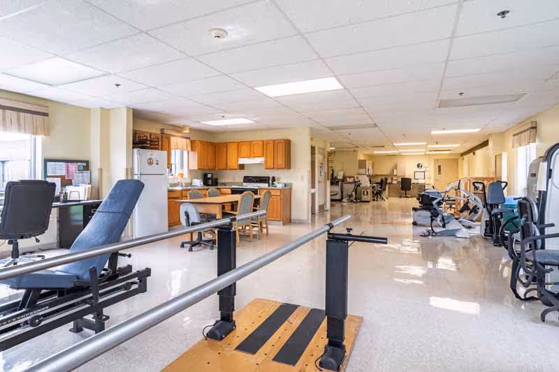 A spacious room in a senior living facility featuring physical therapy equipment including parallel bars and exercise machines. In the background, there is a kitchen area with wooden cabinets, a refrigerator, a stove, and a dining table with chairs. The room has large windows with curtains allowing natural light to enter.