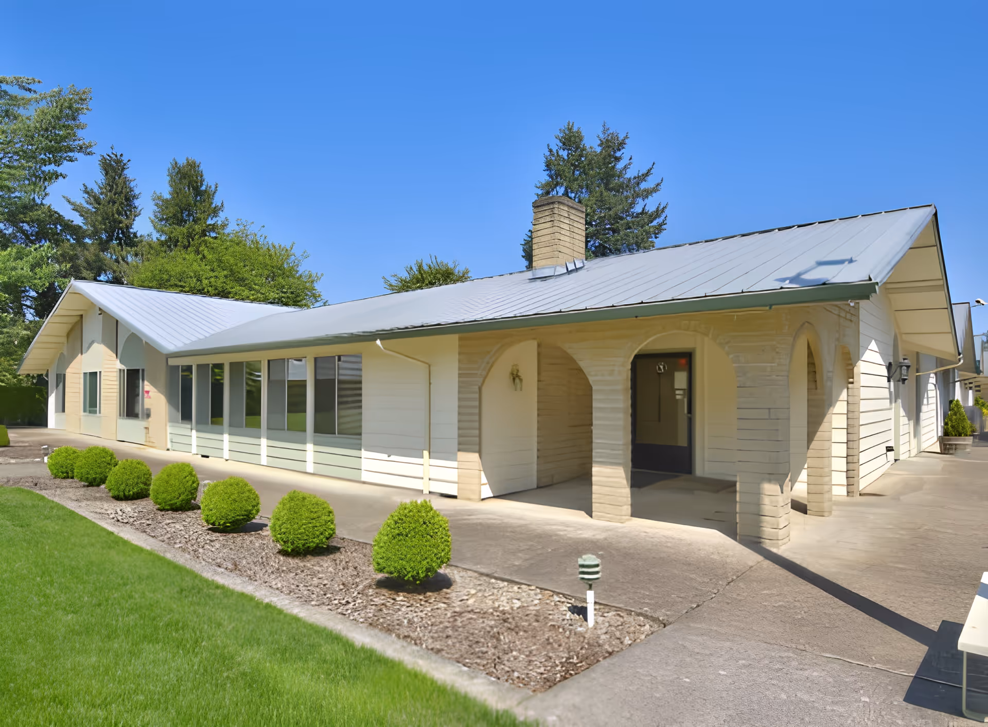 Exterior view of a single-story building with a light-colored facade and a metal roof, featuring arched entryways and large windows. The building is surrounded by neatly trimmed bushes and a well-maintained lawn under a clear blue sky.