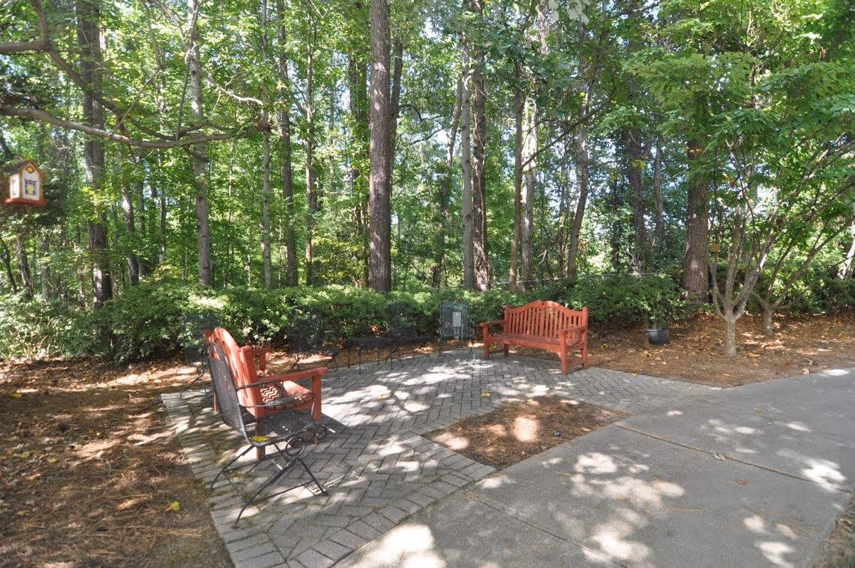 Outdoor seating area with a red wooden bench, two red chairs, and several black metal chairs arranged on a paved patio surrounded by trees and greenery. A birdhouse hangs from a tree branch on the left side.