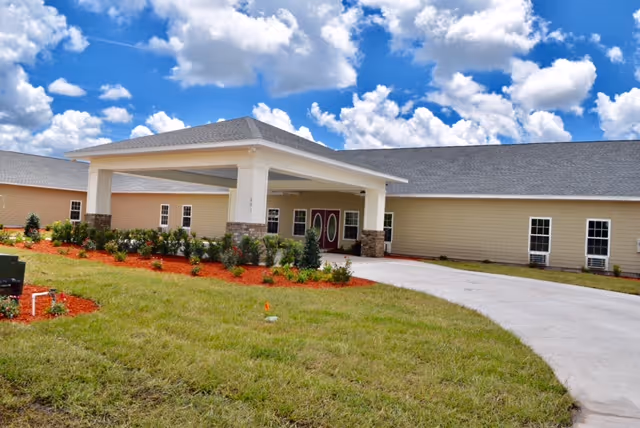Front exterior of a single-story assisted living building with a covered entrance, driveway, and landscaped lawn under a blue sky.