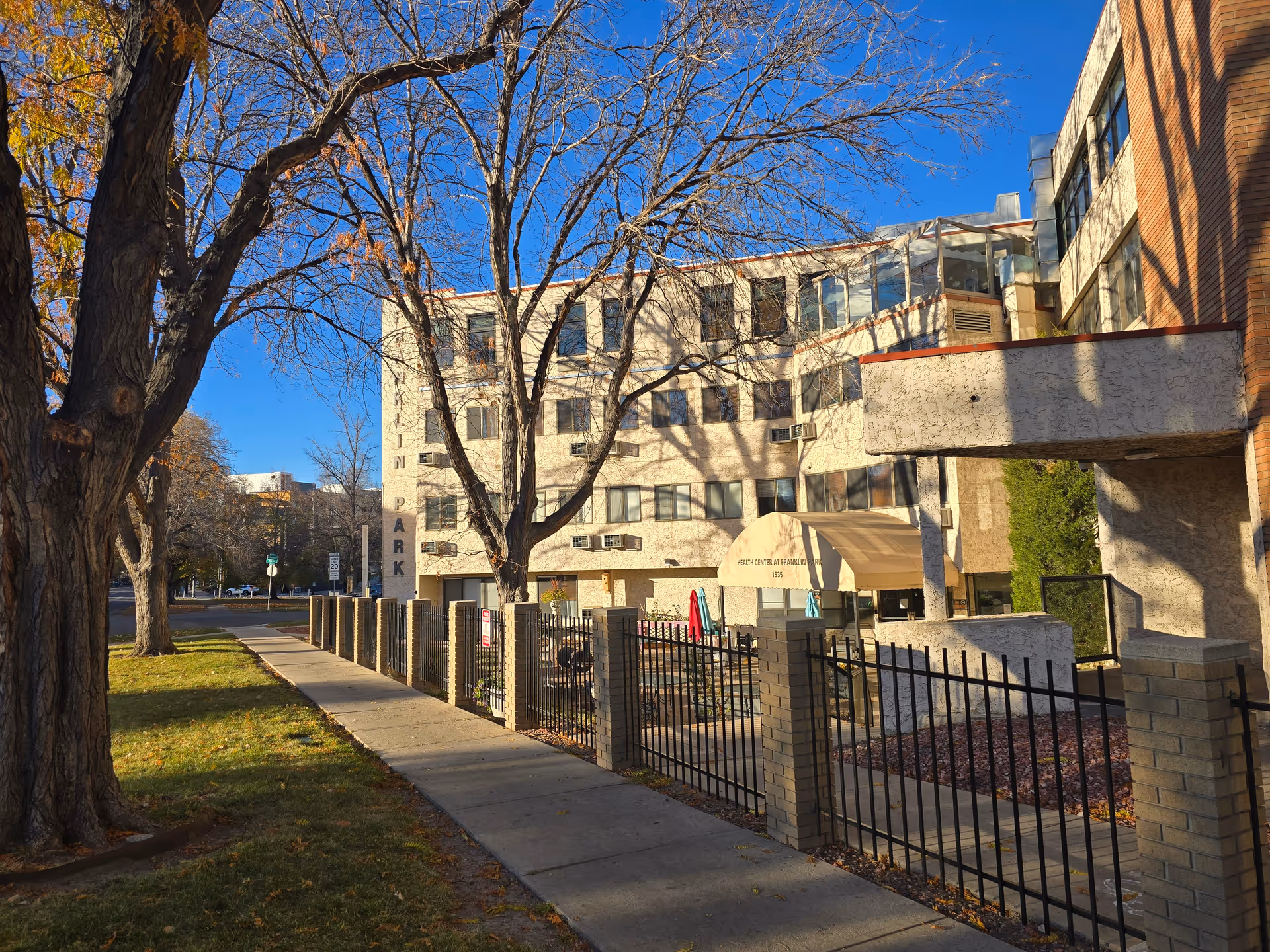 Exterior view of the Health Center at Franklin Park building on a sunny day with clear blue sky. The building is a multi-story structure with numerous windows and air conditioning units. There is a sidewalk along the front with a black metal fence and brick pillars. Leafless trees cast shadows on the building and sidewalk.