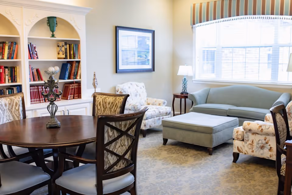 A cozy living room area with a round wooden table surrounded by four upholstered chairs in the foreground. In the background, there is a light blue sofa, a matching ottoman, two floral armchairs, a side table with a lamp, a large window with striped valance, and a built-in white bookshelf filled with books and decorative items.
