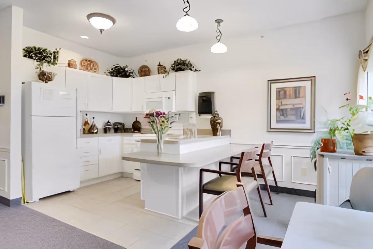 A bright kitchen area with white cabinets, a white refrigerator, and a white countertop island with a vase of flowers. There are three wooden chairs with cushions around the island. The room has decorative plants and framed artwork on the wall, with a window letting in natural light.