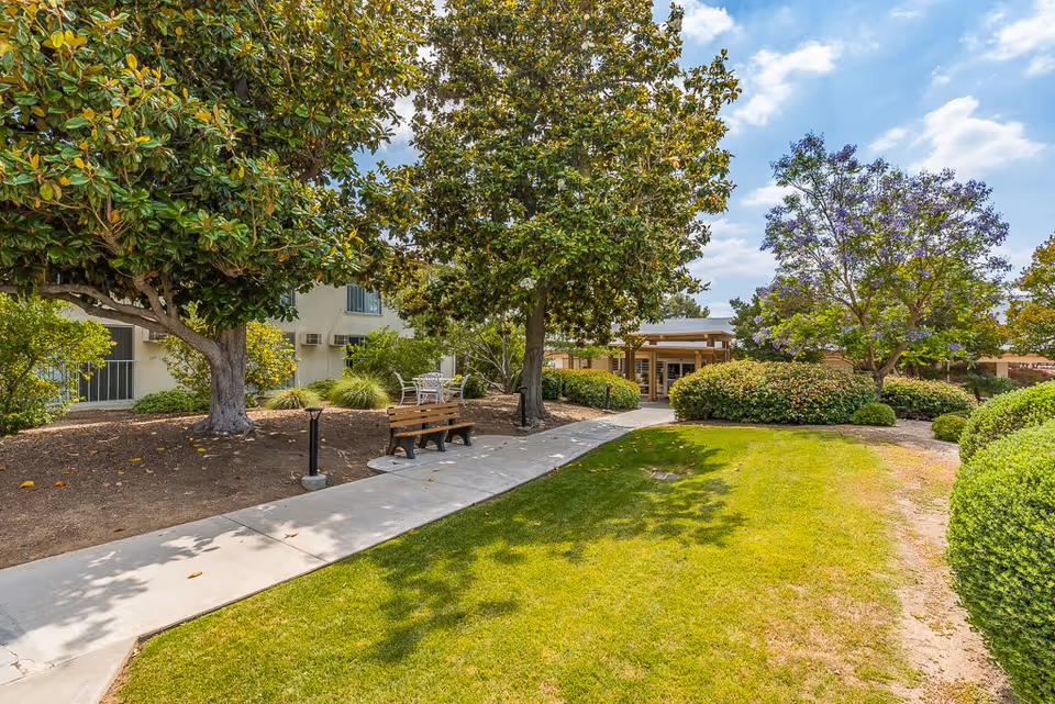 A landscaped outdoor garden area at Sun City Gardens featuring a concrete walkway, a wooden bench, green grass, various trees including a magnolia and a jacaranda with purple flowers, and bushes. The building with windows and air conditioning units is visible in the background under a partly cloudy sky.