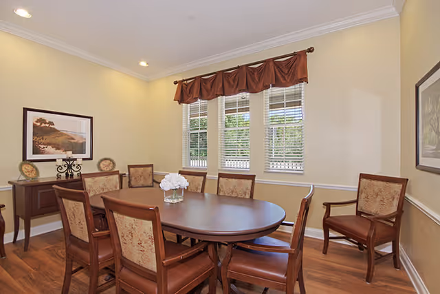 A dining room with a wooden oval table surrounded by eight upholstered chairs. The room has beige walls, a window with white blinds and a brown valance, two framed pictures on the walls, and a wooden side table with decorative items. The floor is wooden and the ceiling has recessed lighting.