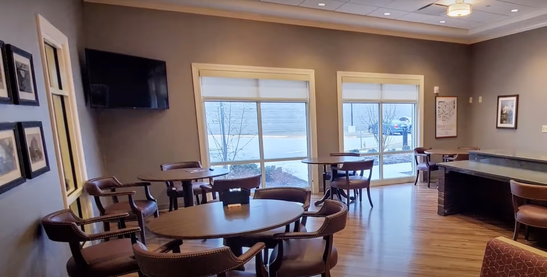 Interior view of a common area in a senior living facility with round wooden tables and brown cushioned chairs arranged around them. The room has large windows with white blinds, framed pictures on the walls, a flat-screen TV mounted on one wall, and a wooden floor. There is a counter or bar area on the right side of the image.