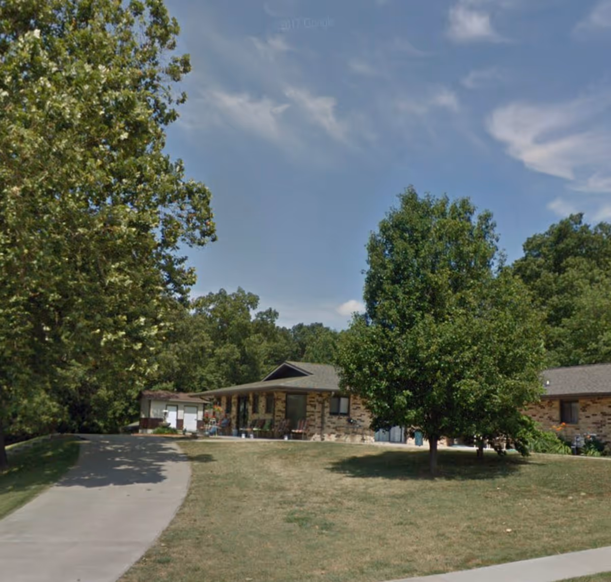 Single-story brick care facility with a driveway, lawn and trees under a blue sky.