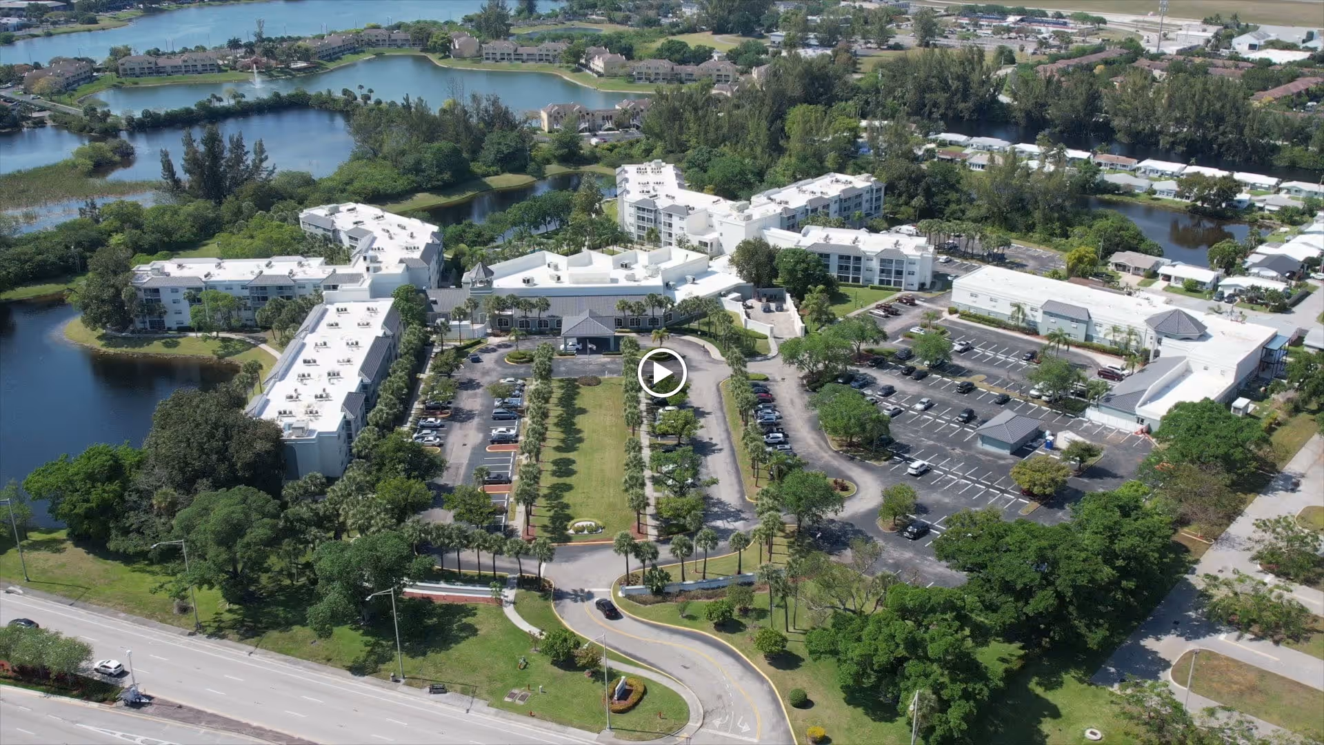 Aerial view of Oasis Living Quarters senior living facility showing multiple white buildings surrounded by trees, parking lots with cars, green lawns, and water bodies in the background.