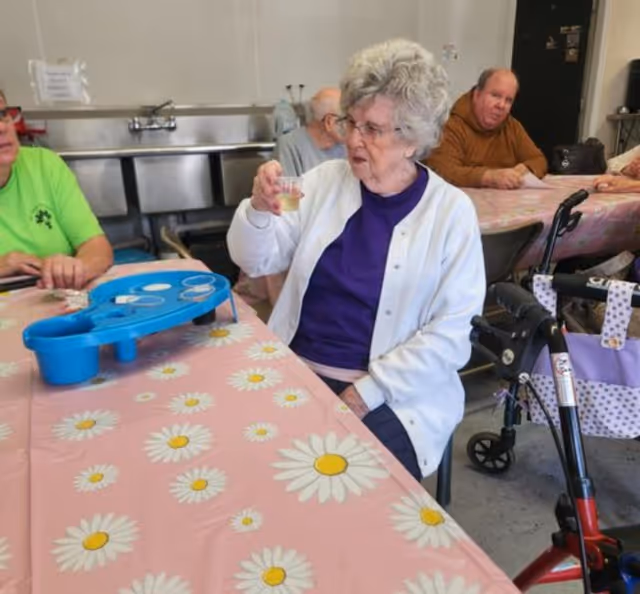 Elderly individuals sitting around a table covered with a pink tablecloth with white and yellow daisies. One elderly woman in a white cardigan and purple shirt is holding a small cup, possibly drinking. There is a blue game or activity board on the table. A walker is visible next to the woman. The setting appears to be a communal indoor space with a stainless steel sink in the background.