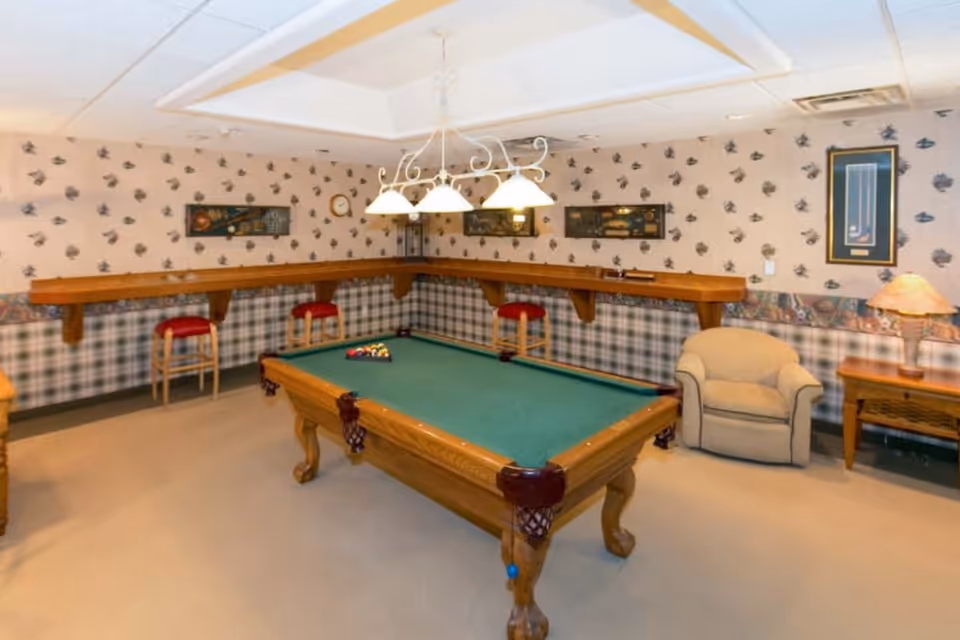A recreational room with a pool table in the center, surrounded by a beige carpet. The walls are decorated with patterned wallpaper featuring small motifs and a plaid lower border. There is a wooden counter with three red cushioned stools along one wall, a beige armchair next to a wooden side table with a lamp, and framed artwork hanging on the walls. A white chandelier with four lights hangs above the pool table.