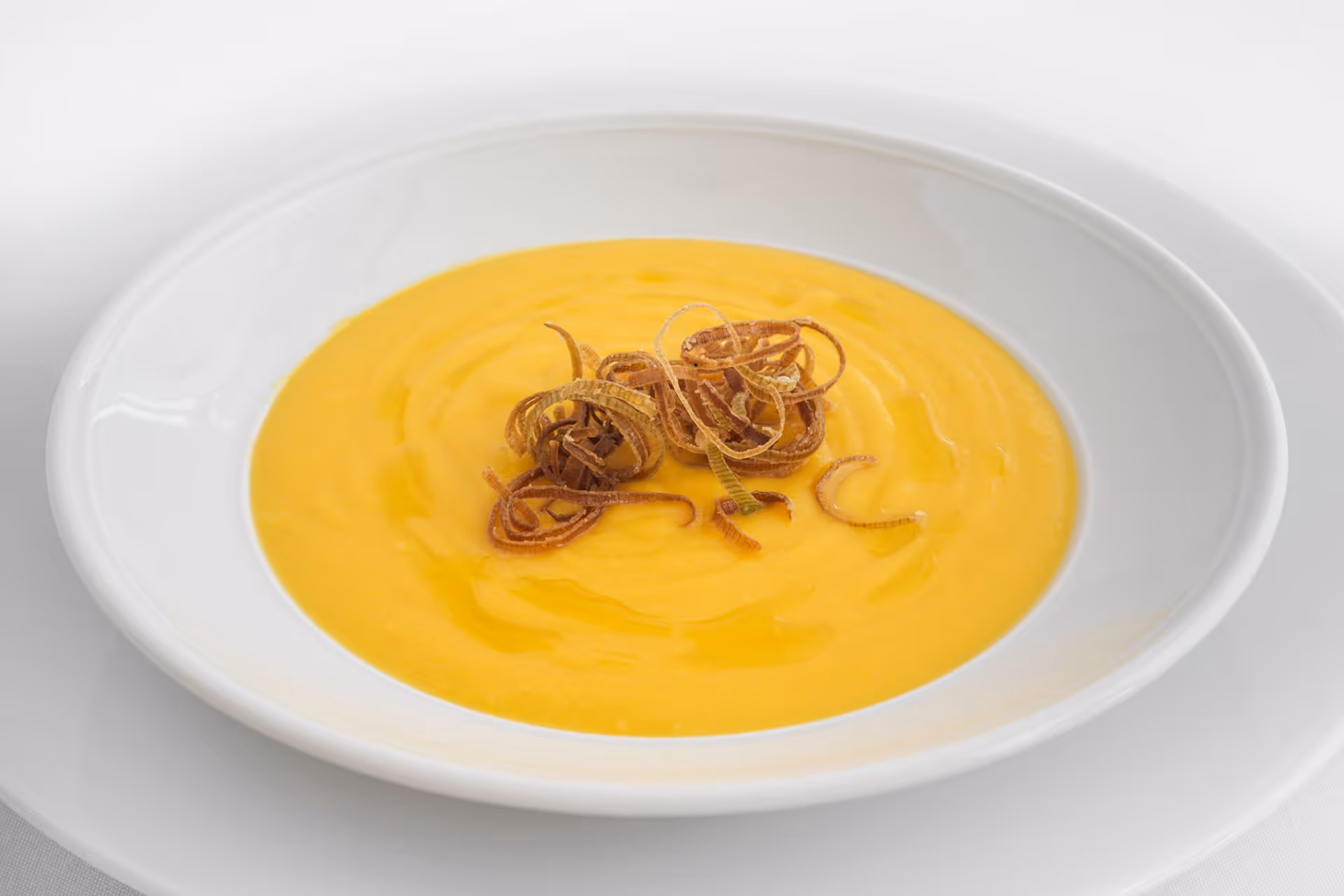 A white bowl filled with creamy yellow soup, garnished with thin, crispy fried onion strips on top, placed on a white plate.