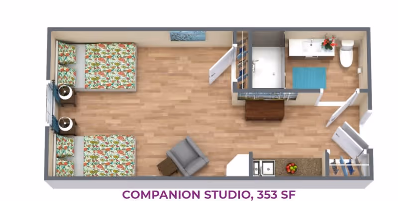 Top-down floor plan of a companion studio apartment featuring two beds with floral bedding, two nightstands, a single armchair with a small table, a bathroom with a shower, sink, and toilet, and a kitchenette area with a sink and countertop. The floor is wood, and there is a closet near the entrance.