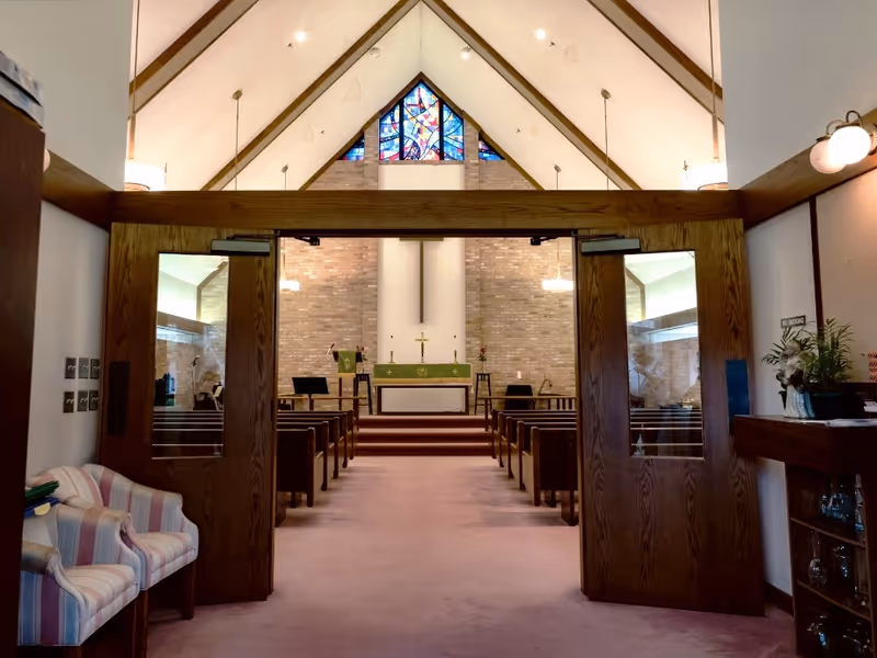 Interior view of a chapel with wooden double doors open, showing rows of wooden pews leading to an altar with a cross and religious items. The chapel has a high vaulted ceiling with stained glass windows at the far end and soft lighting.