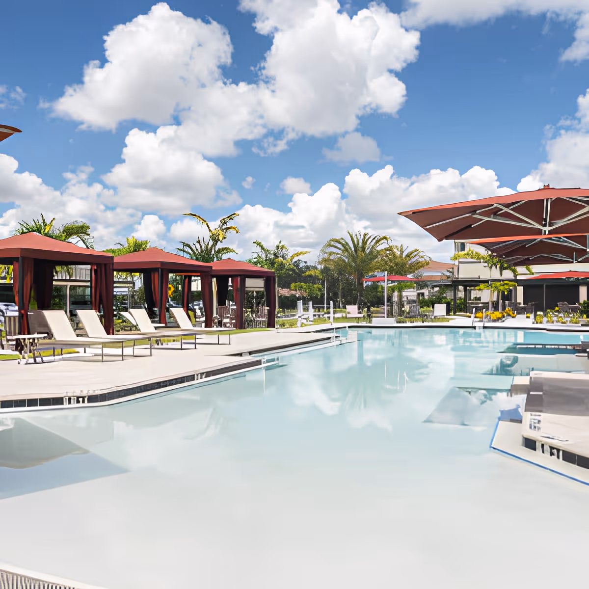Outdoor swimming pool area with several lounge chairs and cabanas with red canopies, large red umbrellas, palm trees, and a bright blue sky with scattered white clouds.