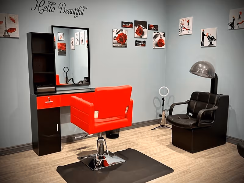Interior salon corner with a red styling chair and mirror at a black vanity, and a black hooded dryer chair against pale blue walls decorated with artwork.